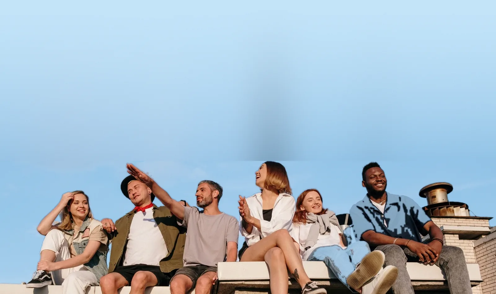 Six friends sit together on a sunny rooftop ledge under a clear blue sky, smiling and laughing