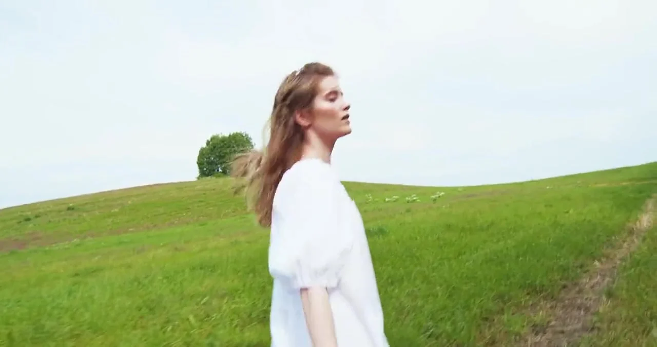 A smiling woman in a white dress runs up a sunny green hill under a bright sky