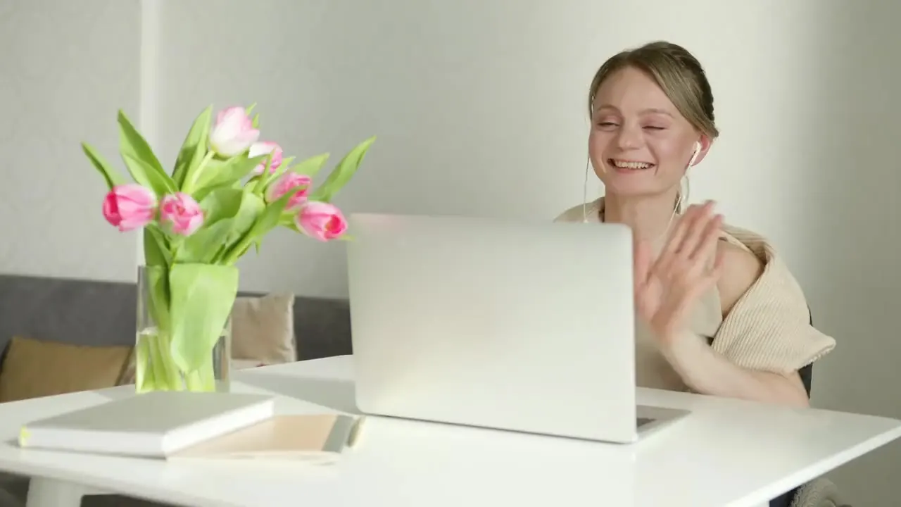A woman at a bright white desk with pink tulips smiles and waves during a video call